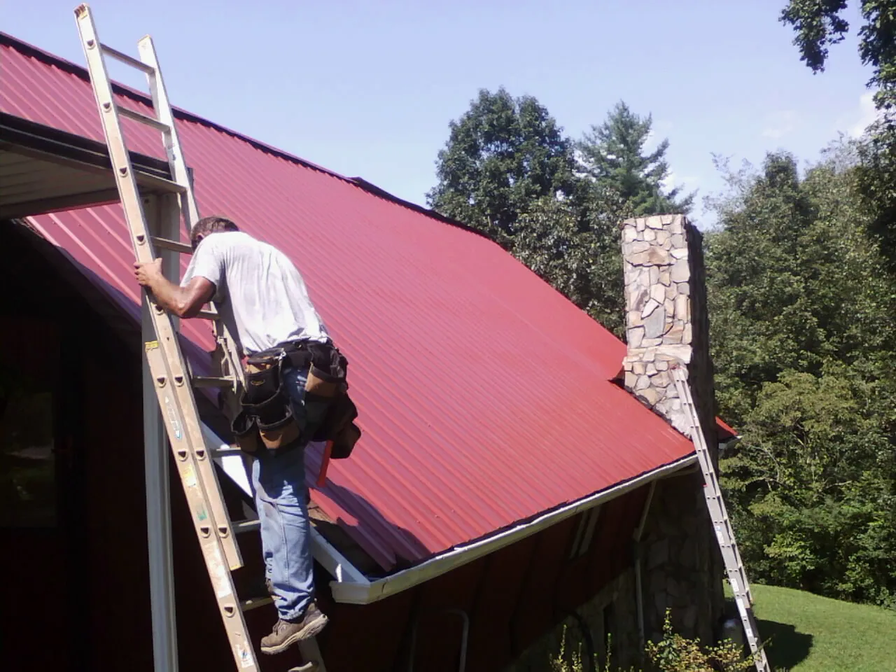 Roofer on a ladder beside a red metal roof and stone chimney — Soffit & Fascia Repair by Roofing Brothers in Cobbs Creek